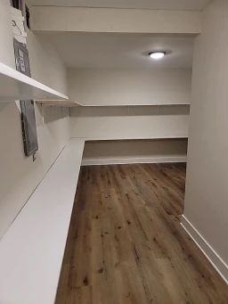 Empty pantry with white shelves and a bench. Light wood flooring.