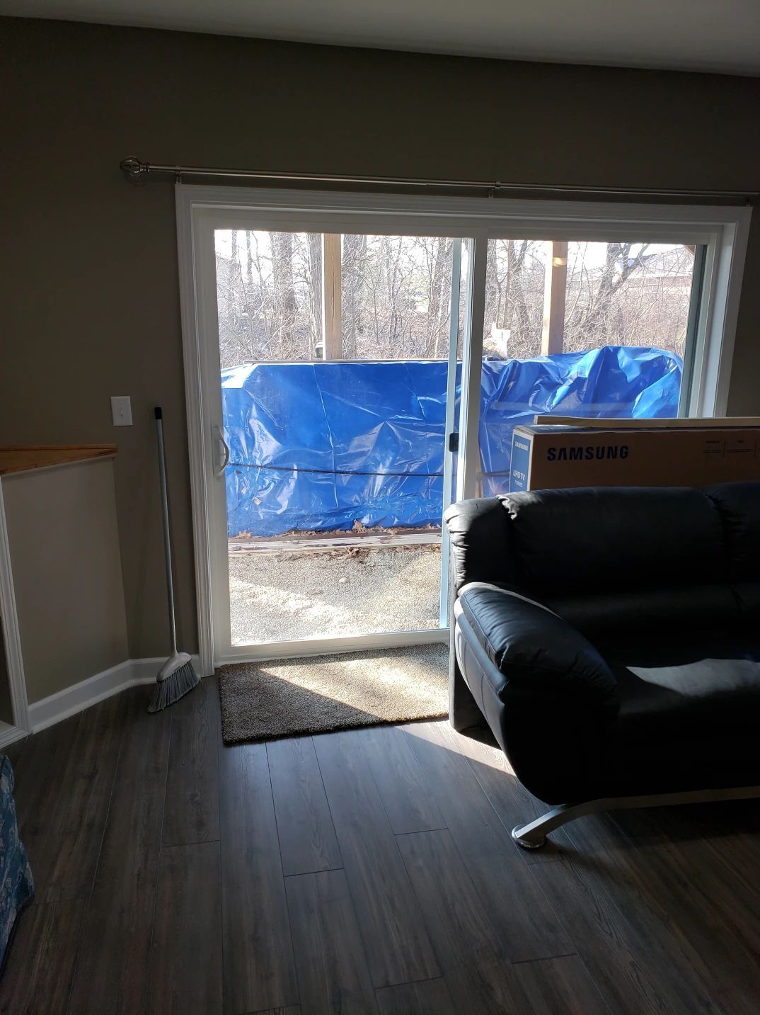 Living room with sliding glass door partially covered by a blue tarp. Sunlight streams in. A couch and a Yamaha box are visible.