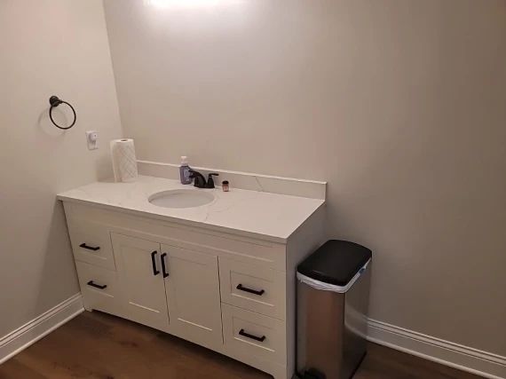 White bathroom vanity with sink, black hardware, paper towels, and trash can.