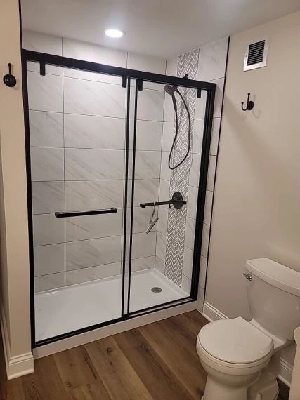 Bathroom with a black-framed shower, white and gray tile, and a toilet. The floor is wood-look.