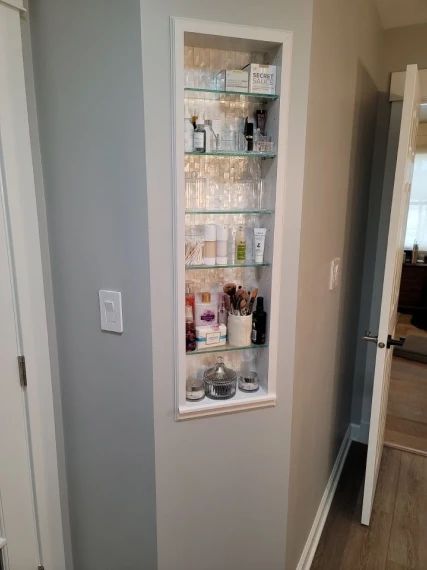 White-framed recessed shelving unit in a hallway, filled with cosmetic products. The walls are gray and beige.