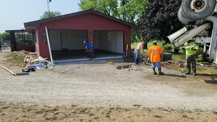 Construction workers pouring concrete into a garage. A cement truck is on the right. Red garage with two bays.