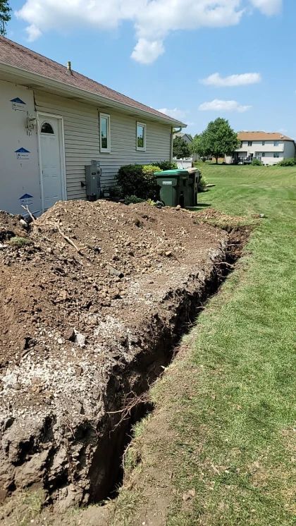 Trench dug along a house foundation, dirt pile beside it, green lawn and residential area in background.