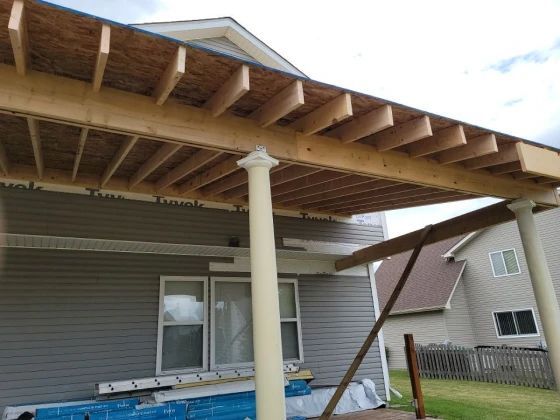 Construction of a deck roof, showing wooden beams, columns, and house siding.
