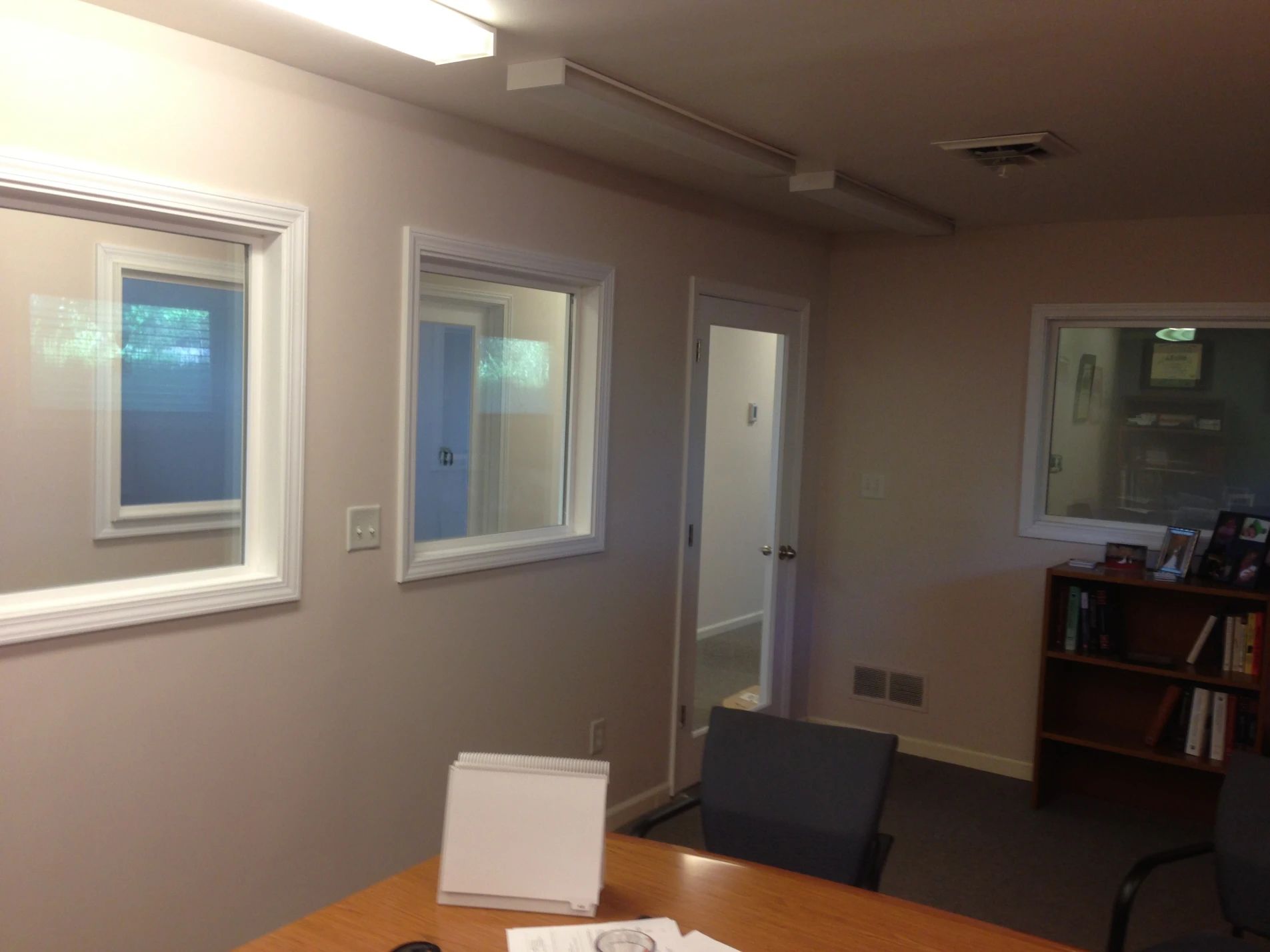 Office interior with windows, desk, bookshelf, and a door to another room. Neutral colors.