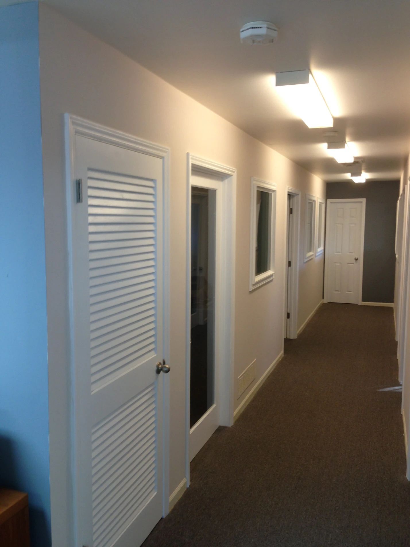 Hallway with doors, windows, and brown carpet, lit by overhead lights.