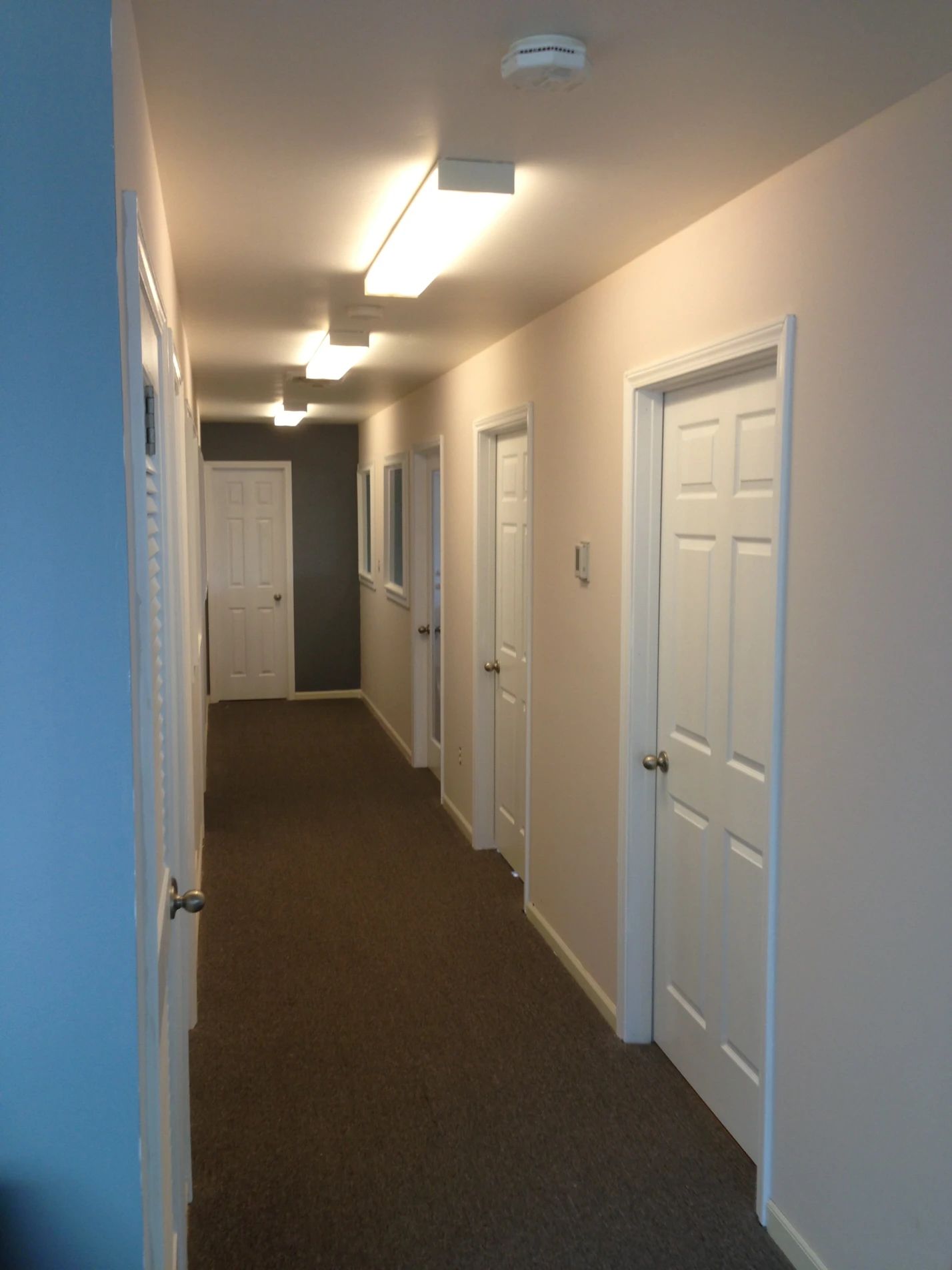 Long hallway with multiple doors, neutral walls, brown carpet, and ceiling lights.