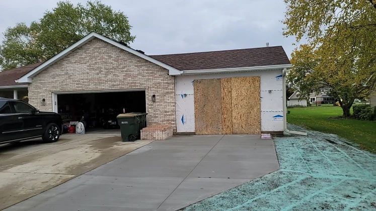 Exterior of a house with two garage doors, one open, the other boarded up with plywood.