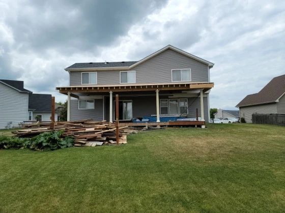 Backyard of a two-story house with a partially constructed deck. Lumber and debris are in the foreground. Overcast sky.