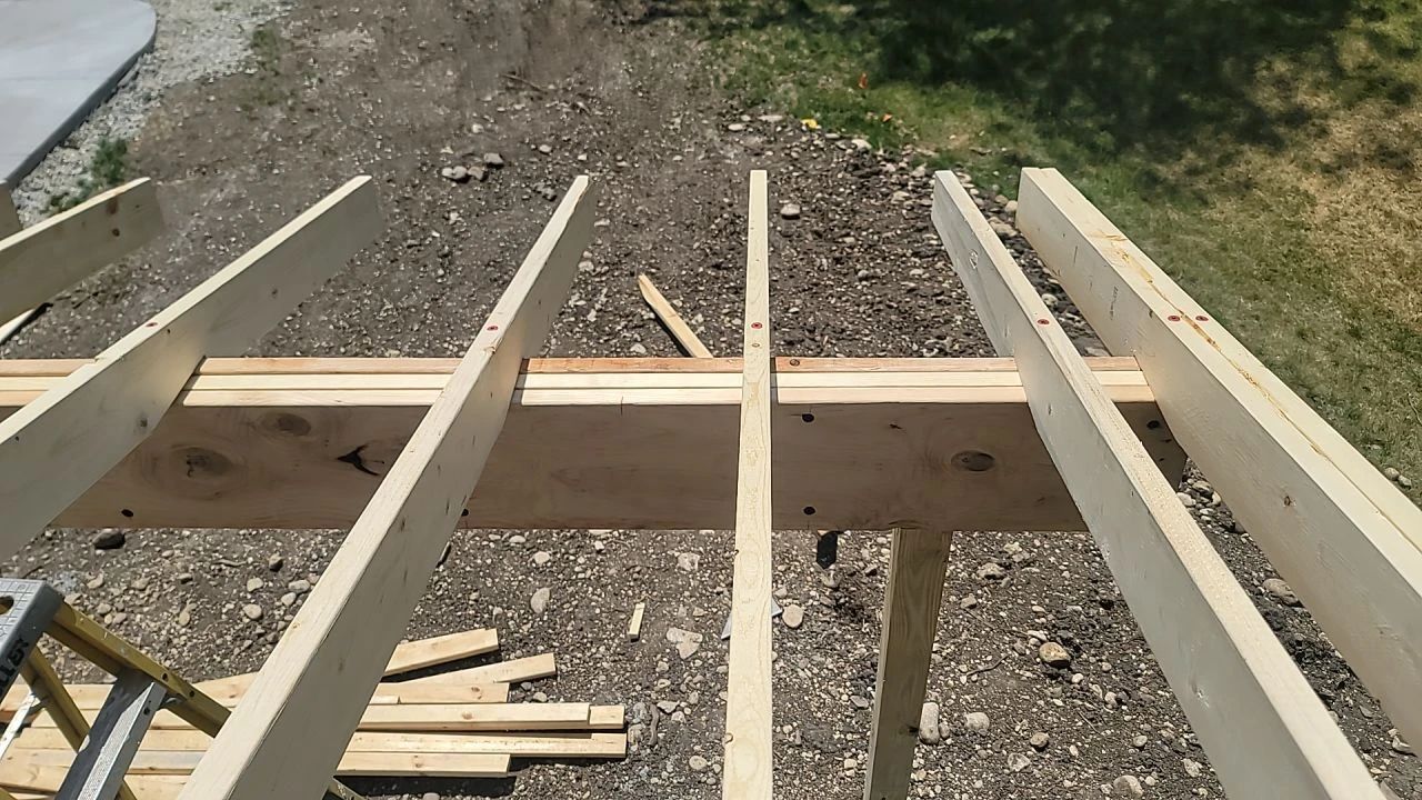 Overhead view of wooden roof framing in progress, with visible rafters and supports against a gravel ground.