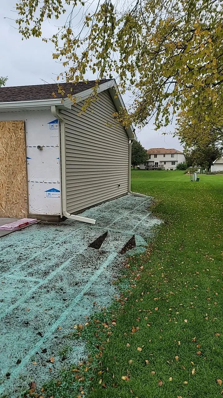 A garage with damaged siding, plywood covering a missing section, and debris on the ground.