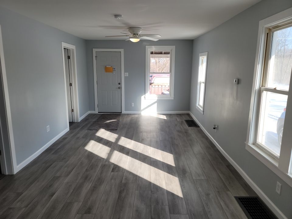 Empty living room with gray walls, wood-look flooring, and windows letting in sunlight.