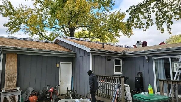 A person on the ground watches roofers working on a house with a gray exterior, under a tree.