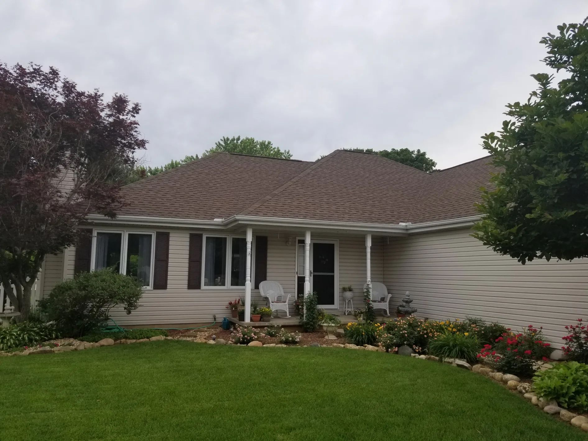 Tan house with brown roof, white porch, and green lawn. Landscaping with flowers and trees.