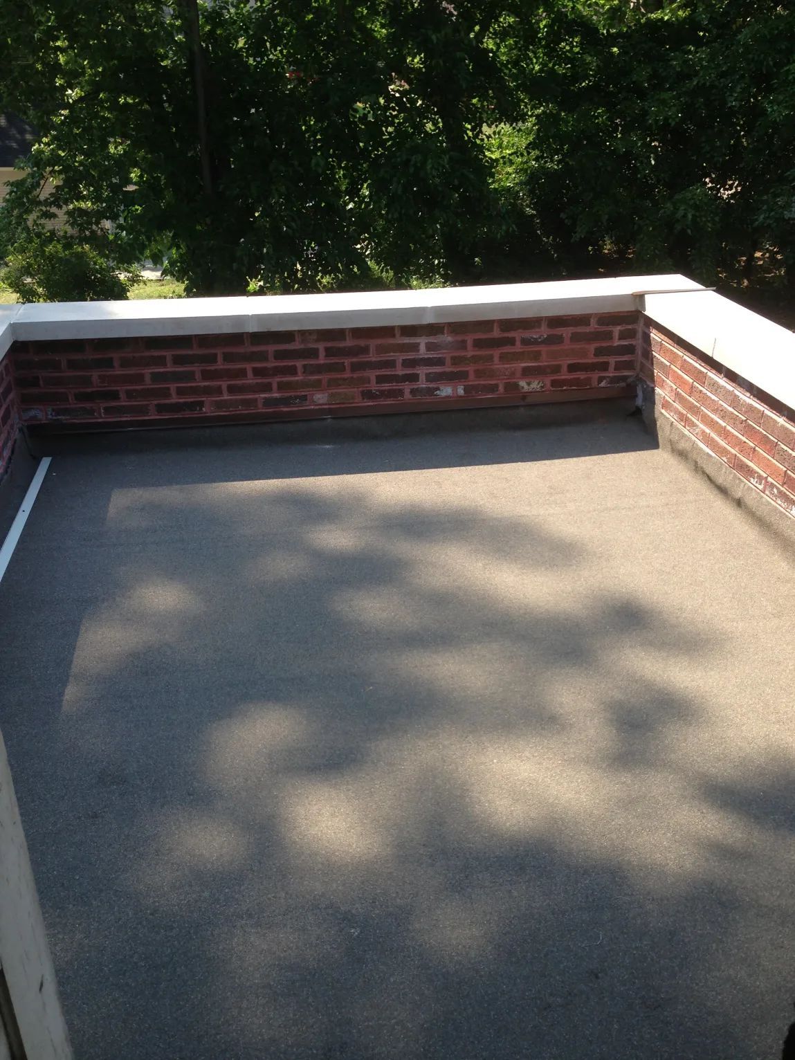 Flat rooftop with brick wall, surrounded by white trim. Shadows cast by nearby trees.