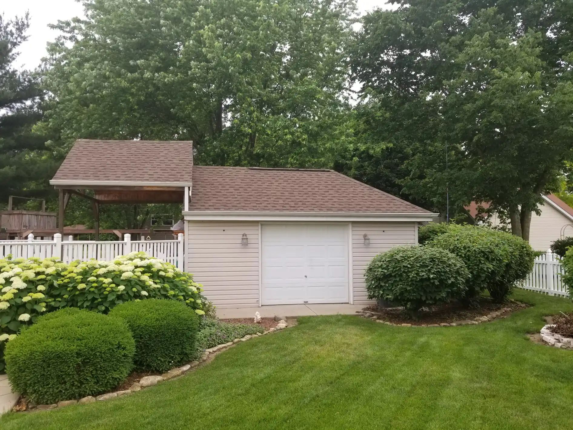 Garage with weathered roof and white door, surrounded by green shrubs, lawn, and fence.