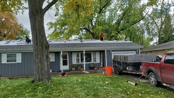 Roofers on a gray house roof, with a trailer and truck in the yard, autumn trees.