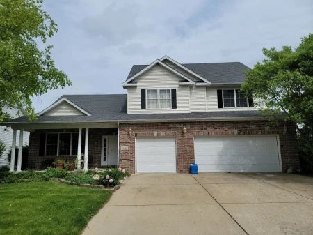 Two-story brick house with a black roof and attached garage. A porch with white columns is in front.