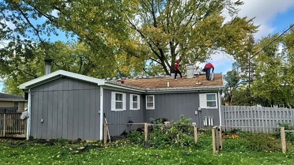 Two workers on a gray house roof removing shingles, surrounded by green trees and a fence.