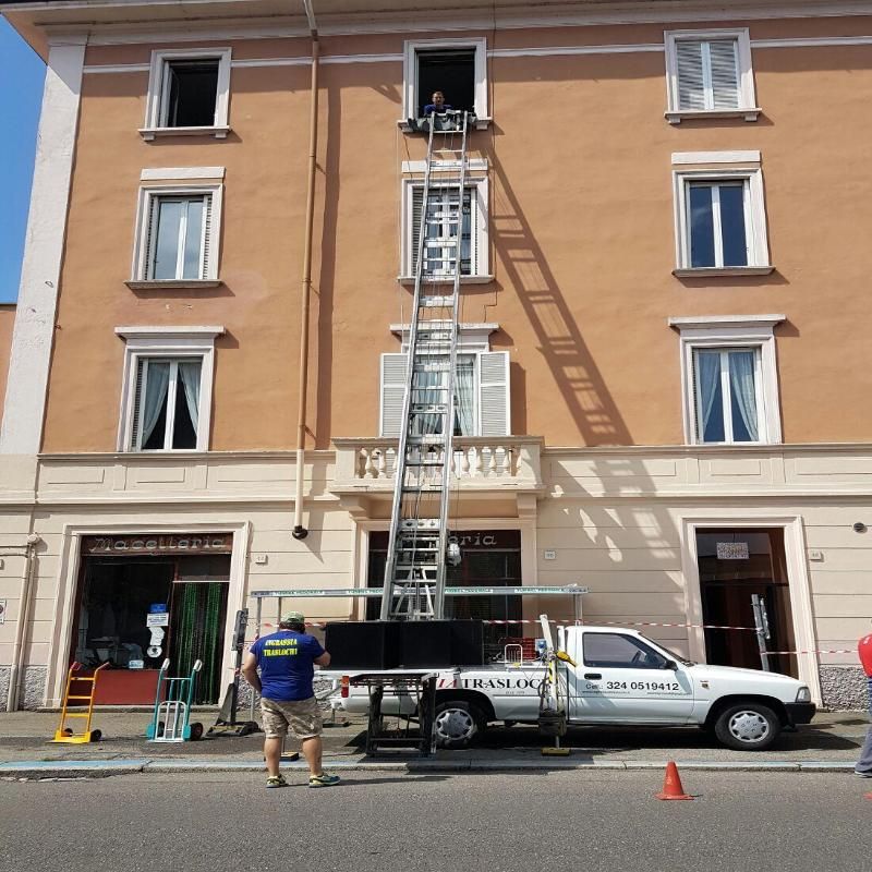 Camion in movimento con un elevatore appoggiato a un edificio; un operaio osserva gli oggetti che vengono spostati dentro e fuori da una finestra.