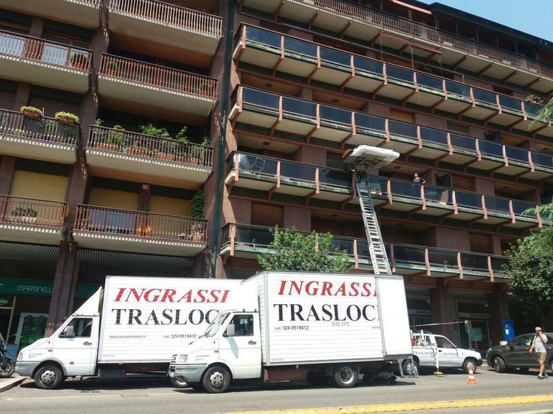 Due camion per traslochi con piattaforma elevatrice, parcheggiati vicino a un condominio. Sui camion è stampata la scritta 