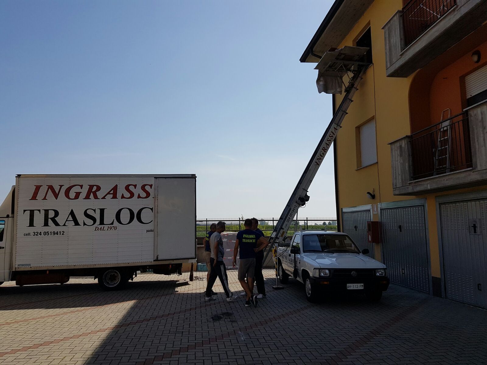 Un camion con piattaforma elevatrice scarica mobili dalla finestra di un edificio. Tre persone sono in piedi nelle vicinanze.