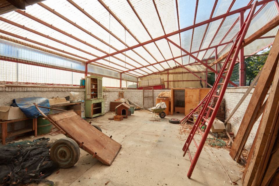 Interno di un magazzino con una scala rossa, assi di legno e vari attrezzi, illuminato dalla luce del sole che filtra attraverso un tetto traslucido.