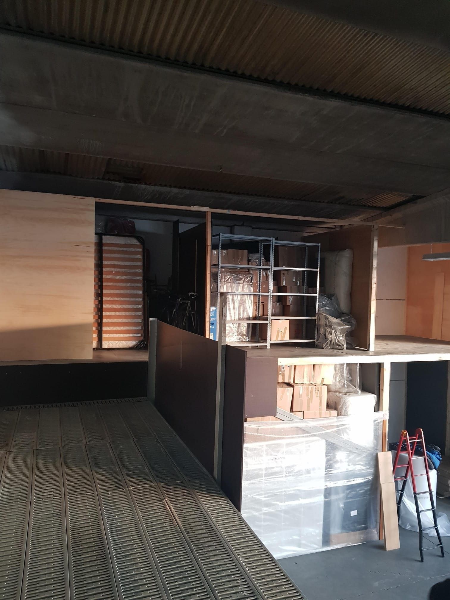 Interno di un magazzino con scaffali, travi di legno, vari oggetti e una scala.