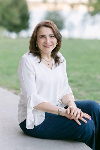 Woman in a white blouse and jeans sitting outdoors on a sidewalk, smiling at the camera.