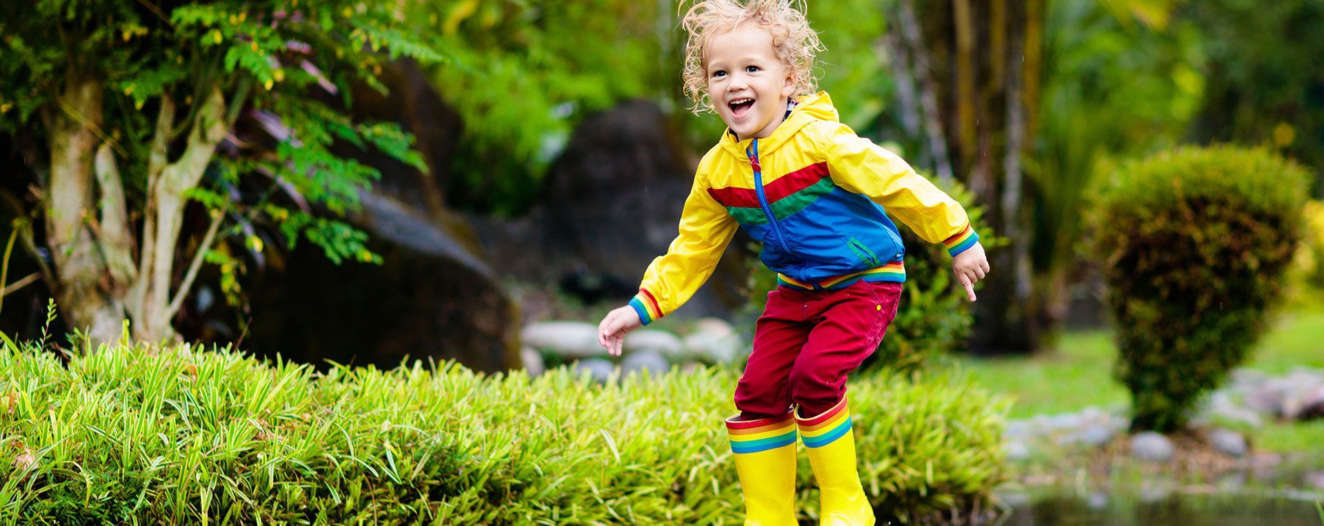 Child in a yellow jacket and red pants running through a green garden, smiling.