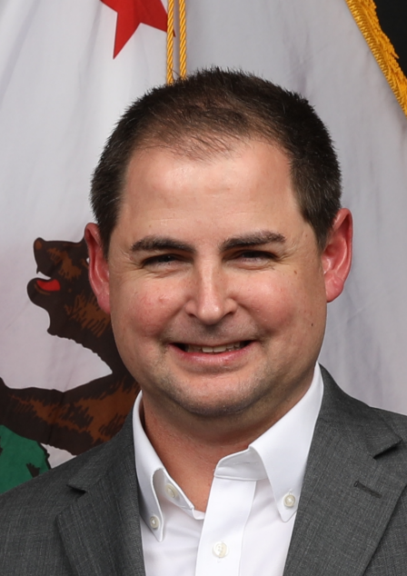 A man in a suit is smiling in front of a california flag