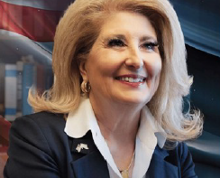 Denice Gary-Pandol Smiling blond woman in a dark suit, white shirt, and gold earrings. A partial American flag and building are in the background.