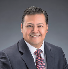 Jack Guerrero Man in suit smiling, with dark hair, and a patterned tie against a gray background.
