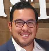 Josh Rodriguez Man with glasses, smiling, wearing a blue blazer and white shirt, set against a wooden background.