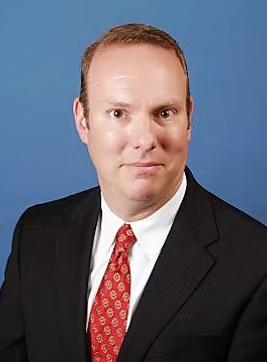 A man in a suit and tie is standing in front of a blue background.