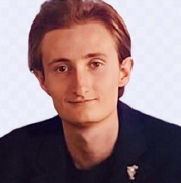 A person with light-brown hair, wearing a dark blazer with a small pin, looking forward against a white background.