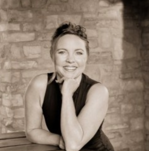 Tracey Coe Woman with dark hair, smiling, resting chin on hand, leaning on a bench near a stone wall.