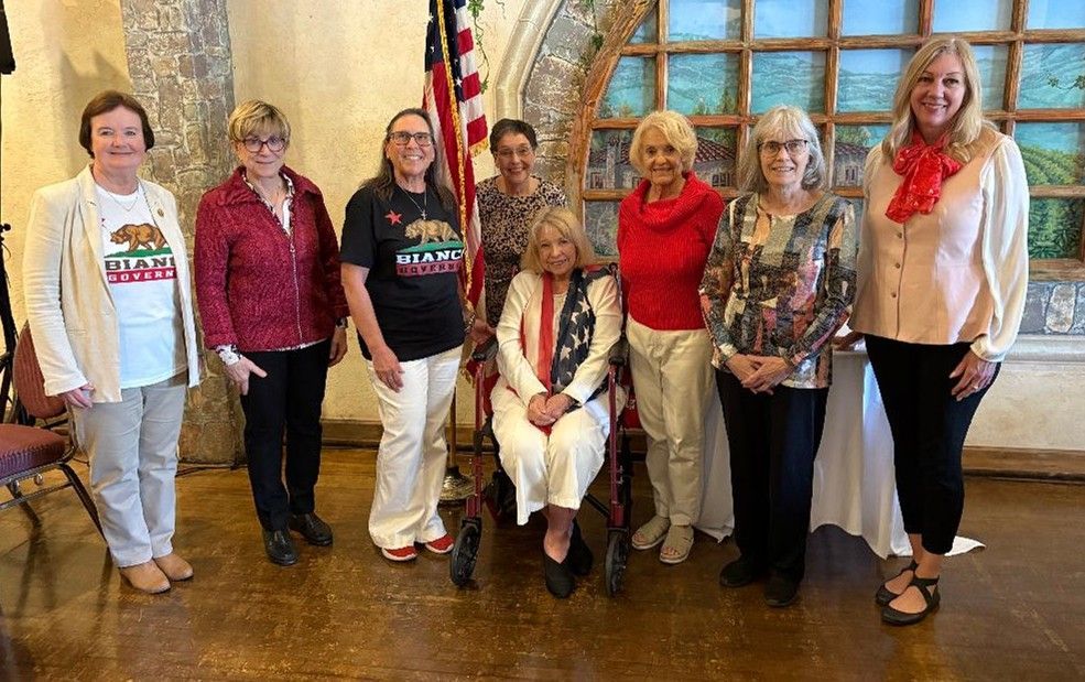 Group of women posing, some with US flag, indoors with arched windows.