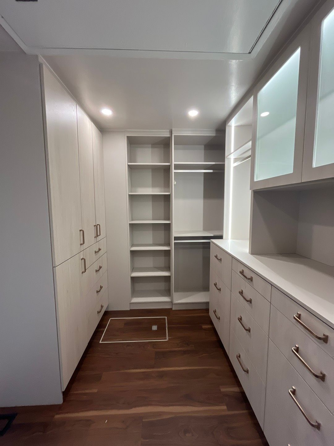 Walk-in closet with light-colored cabinets, shelves, drawers, and wooden floor. LED lighting illuminates the space.