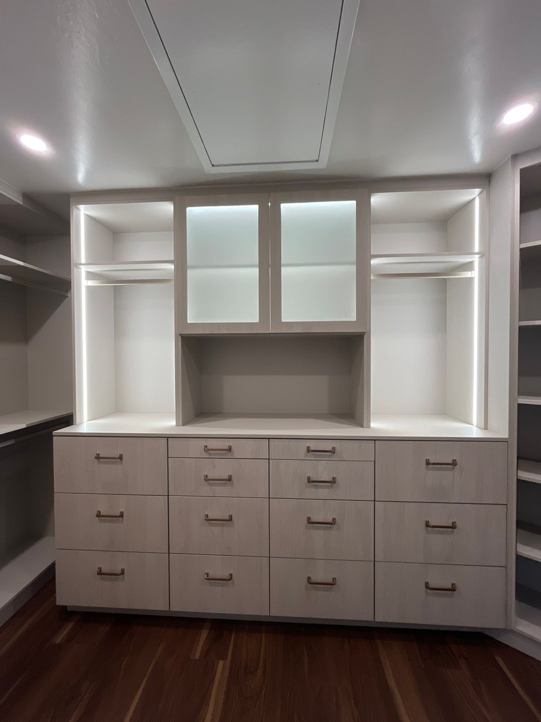 Walk-in closet with white cabinets, drawers, hanging rods, and glass-door upper cabinets, lit by overhead and cabinet lighting.