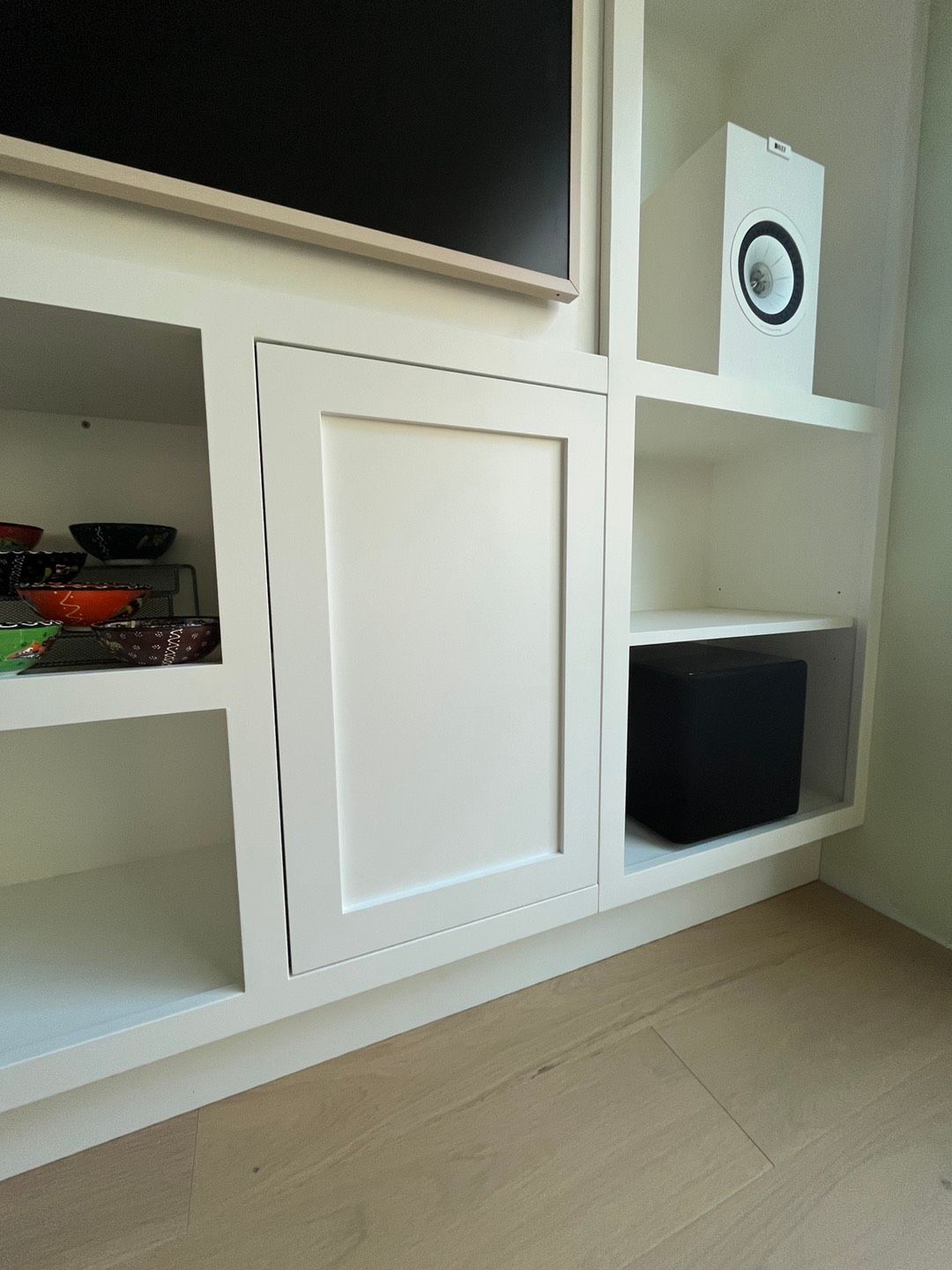 White built-in cabinet with open shelves, a closed door, and a black box. A white speaker sits above.