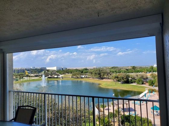 A balcony view overlooking a pond with a fountain, a small patio area, and distant buildings under a sunny, blue sky.