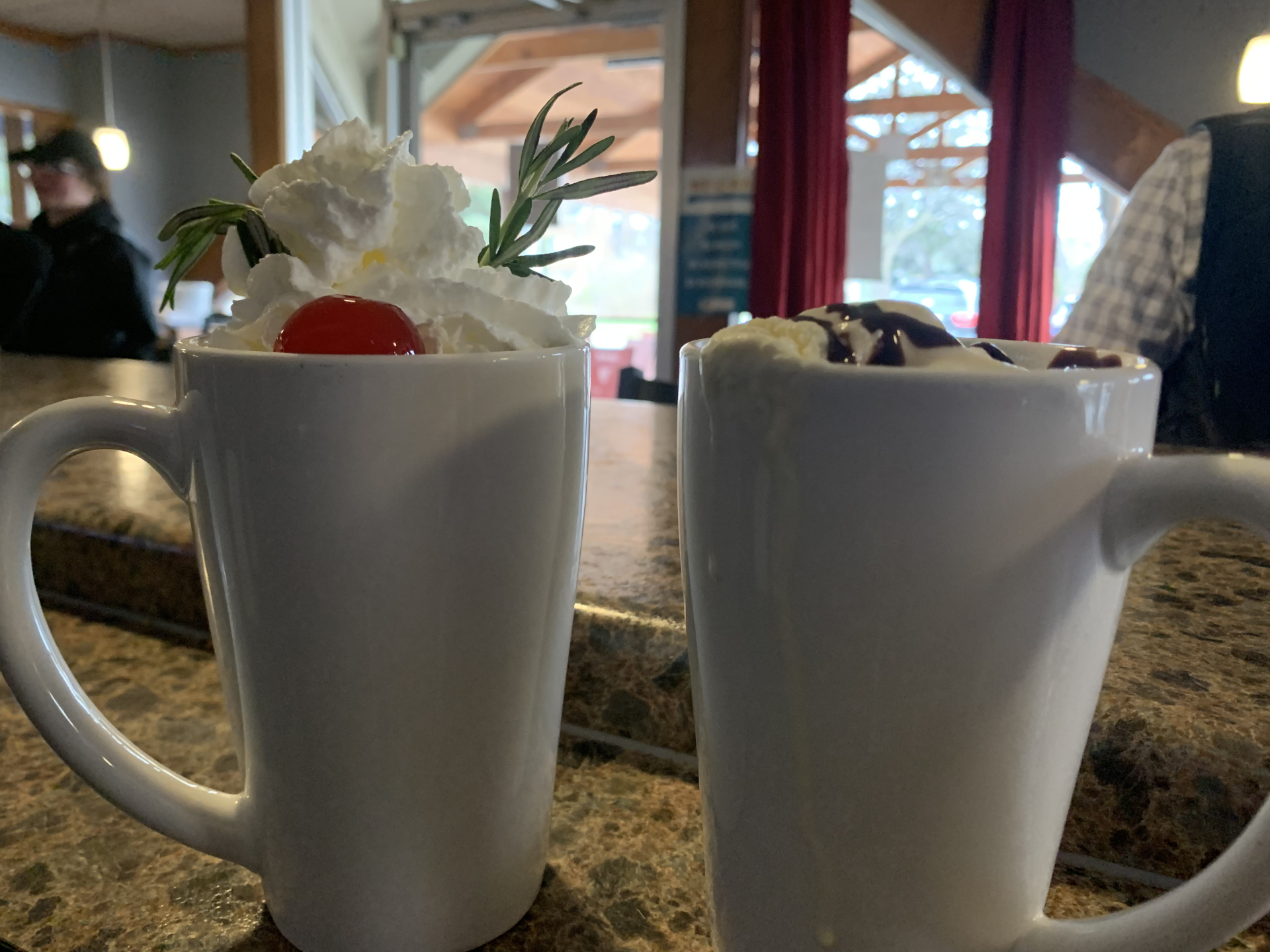 Two mugs of hot chocolate with whipped cream and strawberries on a counter.