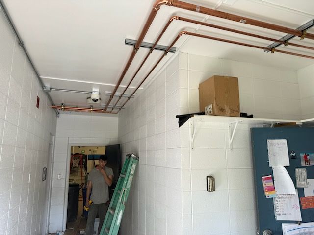 A man with a ladder is standing in a room with copper pipes hanging from the ceiling.