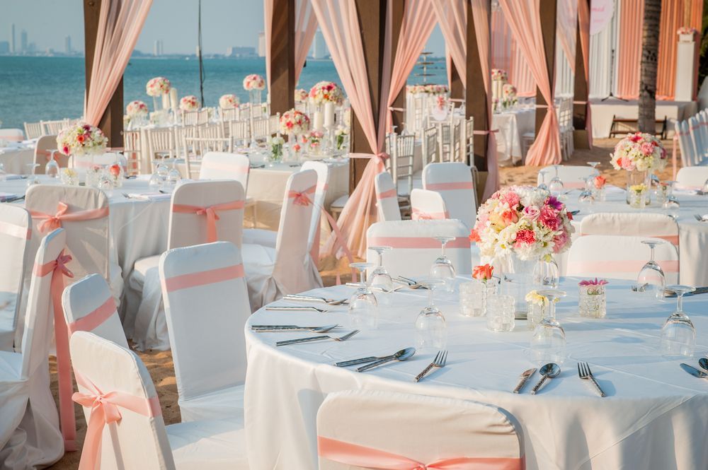 Elegant outdoor wedding reception with white tables, pink chairs, floral centerpieces, and sea views