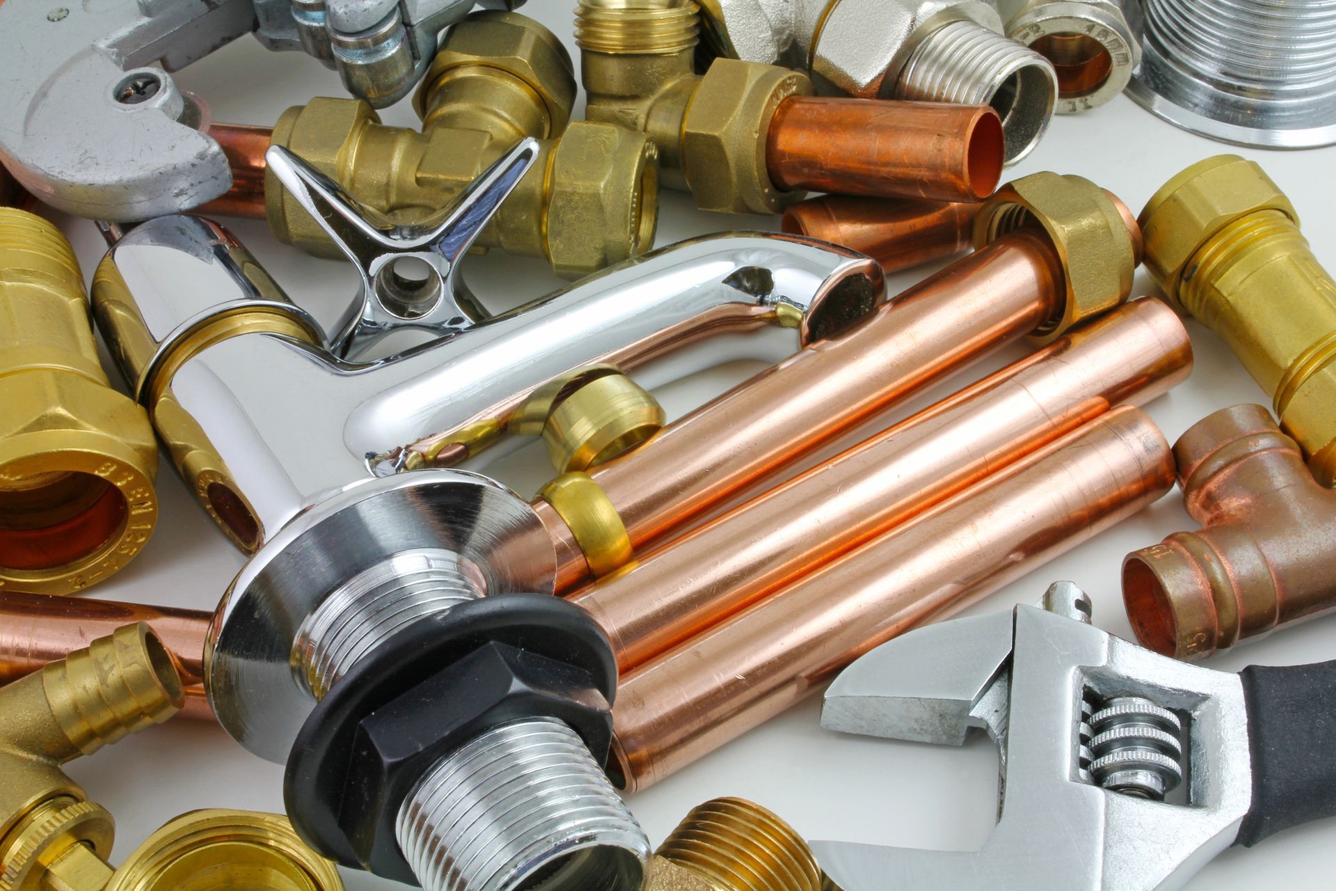 Una collezione di tubi di rame, raccordi idraulici in ottone, un rubinetto cromato e una chiave inglese regolabile su una superficie bianca.