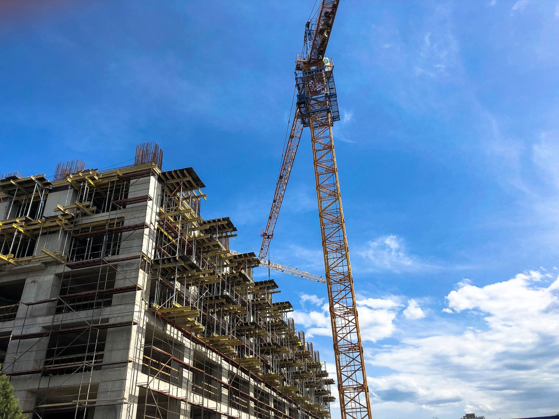 Un'alta gru a torre gialla si erge accanto a un grande edificio a più piani in costruzione, stagliandosi contro un cielo azzurro brillante.