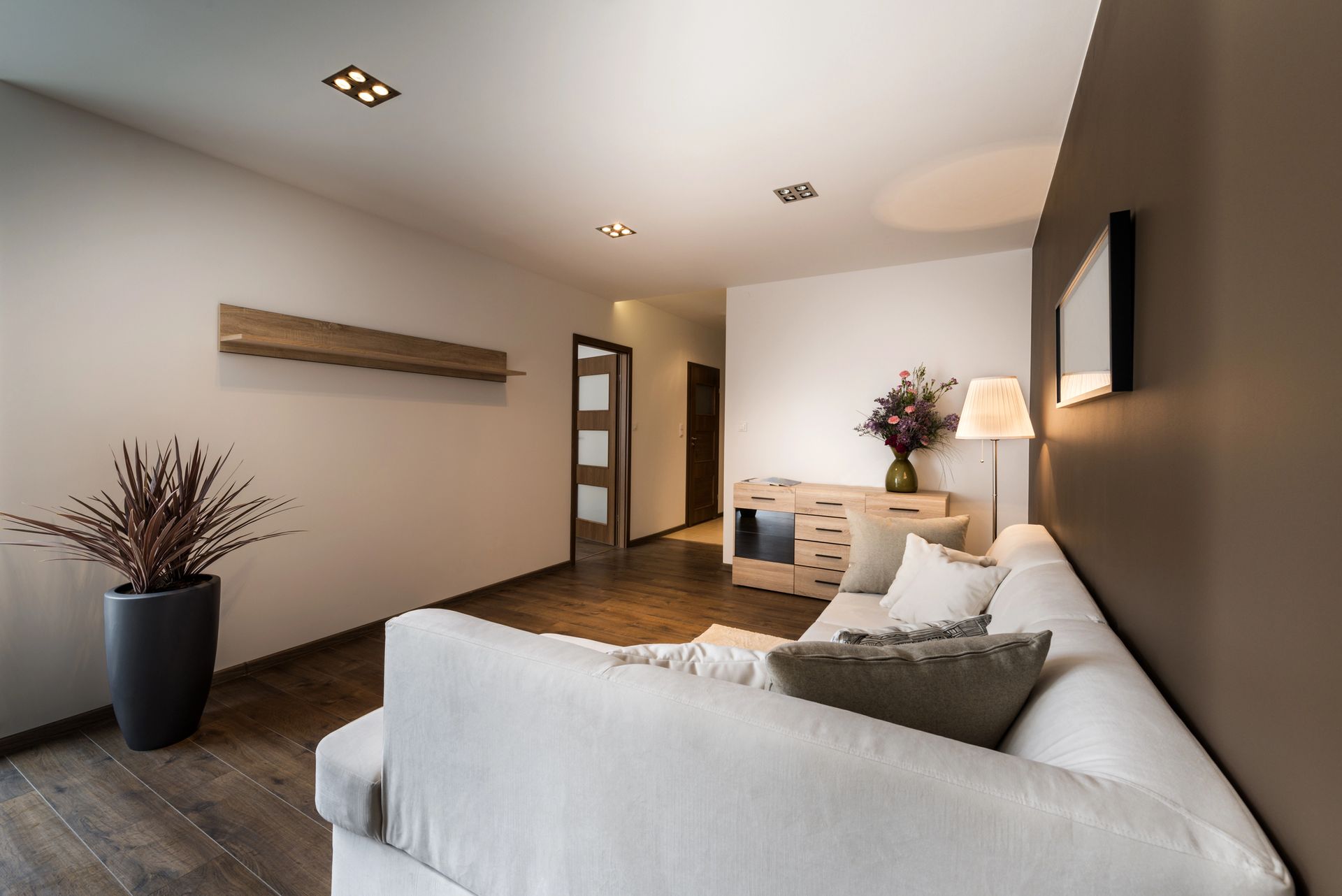 Soggiorno moderno con divano bianco, parete d'accento marrone scuro, mensole in legno e una grande pianta in un vaso grigio scuro.