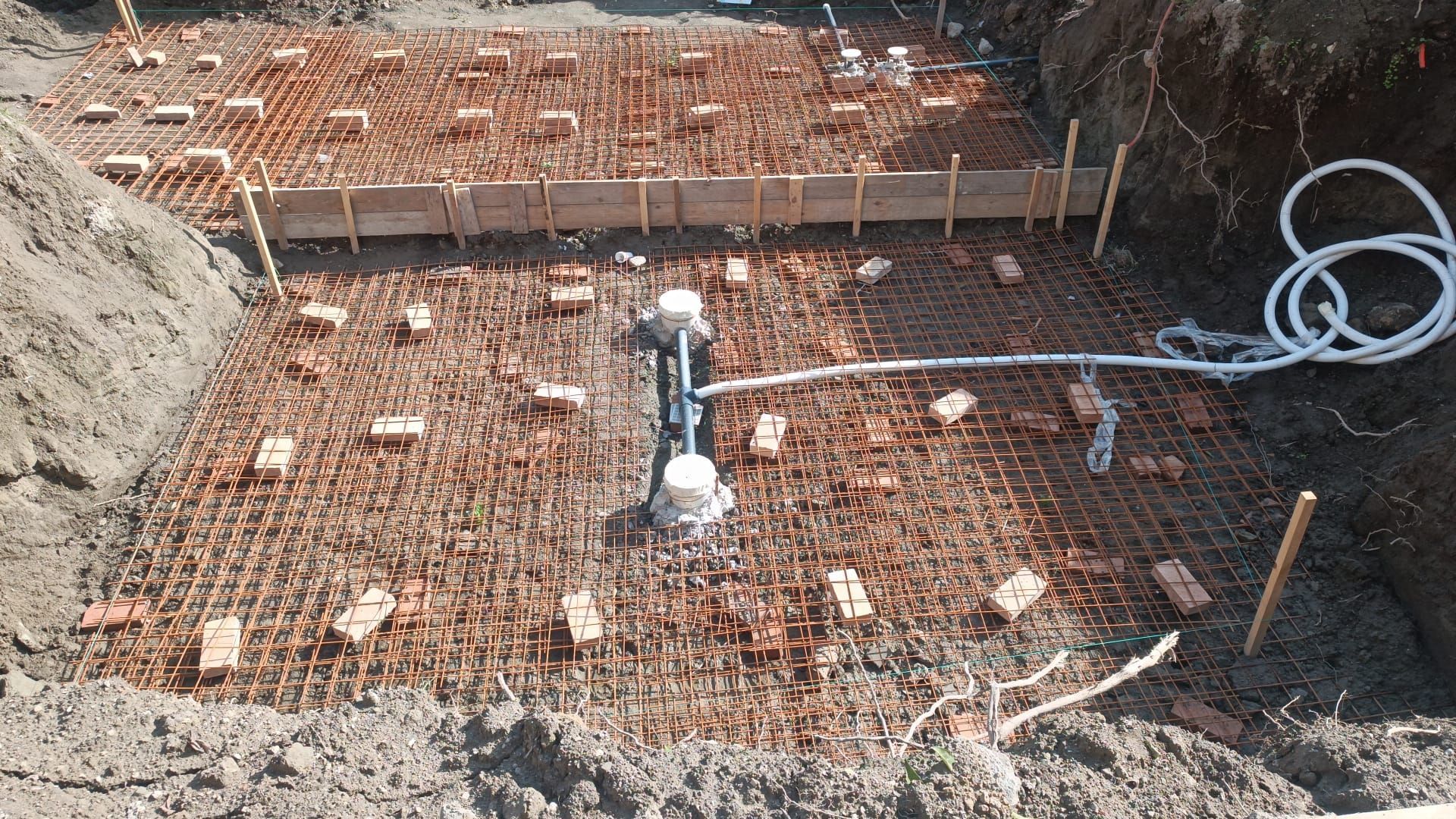 Cantiere edile che mostra la rete di rinforzo in acciaio posata per una fondazione in cemento, con tubature idrauliche a vista.