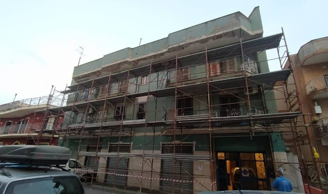 Un edificio verde a più piani, in fase di ristrutturazione esterna, circondato da impalcature metalliche, situato in una via cittadina.